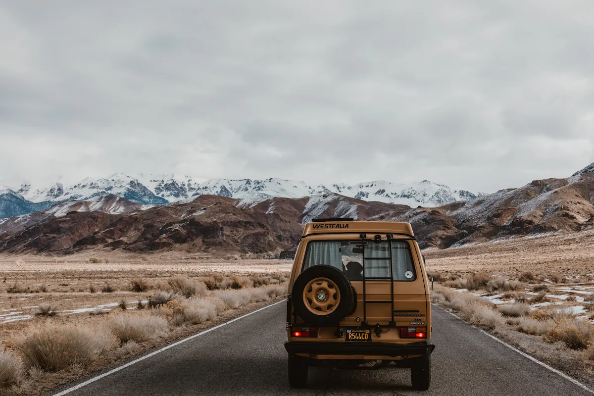 A tan van with a spare tire on the back drives down a deserted road towards snow-covered mountains under a cloudy sky. The barren landscape features dry grass and sparse vegetation on either side of the road.