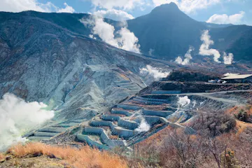 Owakudani Valley A scenic view of a mountainous landscape with terraced slopes and steam rising from multiple locations. The sky is partly cloudy, and the sun casts light over the hazy scene, accentuating the rugged terrain. Sparse vegetation is visible in the foreground.