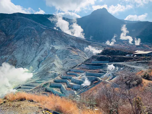 Owakudani Valley A scenic view of a mountainous landscape with terraced slopes and steam rising from multiple locations. The sky is partly cloudy, and the sun casts light over the hazy scene, accentuating the rugged terrain. Sparse vegetation is visible in the foreground.
