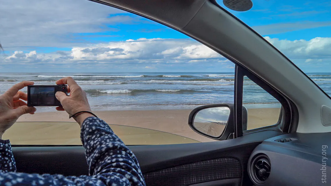 A person seated inside a car is taking a photo of a beach through the open passenger-side window. The sunny beach and ocean waves are seen through the window. The interior of the car, including the dashboard and a side mirror, is visible.