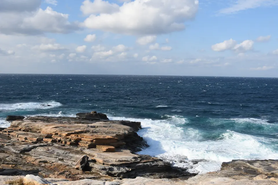 A rocky coastline with waves crashing against the shore under a partly cloudy sky. The horizon line separates the deep blue sea from the sky, creating a tranquil seascape.