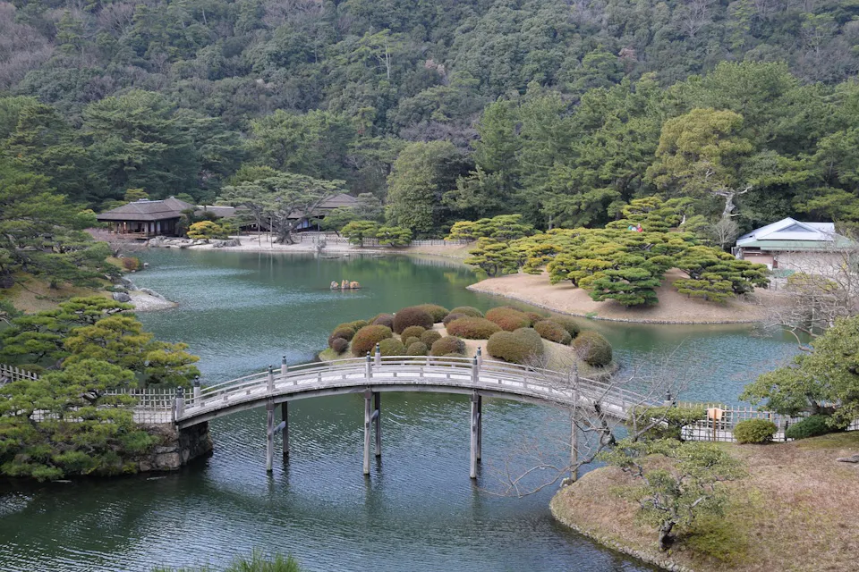 A serene Japanese garden featuring a small lake with a curved wooden bridge. The landscape includes manicured trees, shrubs, and several traditional wooden structures surrounded by lush greenery under a cloudy sky.