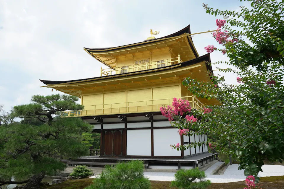 A golden pavilion with traditional Japanese architectural design stands surrounded by lush green trees and pink flowers. The structure features multiple levels and a prominent roof with upward-curving eaves. The lower part of the pavilion is white with wooden doors.