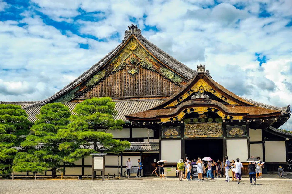 Image of Nijo Castle in Kyoto, Japan, featuring traditional Japanese architecture with an elaborate roof and detailed woodwork. A group of tourists are gathered near the entrance, some holding umbrellas. Pine trees line the front, and the sky is partly cloudy.