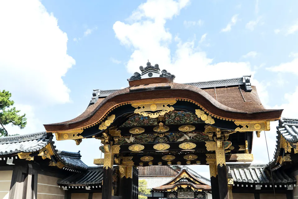A traditional Japanese gate with an intricately decorated roof featuring gold embellishments and detailed carvings. The structure is set against a backdrop of a clear blue sky with a few scattered clouds. Other architectural elements are visible in the background.