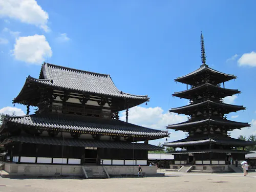 Horyu-ji Temple A sunny day highlighting the ancient wooden structures of Horyu-ji Temple in Japan. The image features a large, traditional main hall on the left and a five-story pagoda on the right, both showcasing classic Japanese architecture against a backdrop of blue sky and scattered clouds.