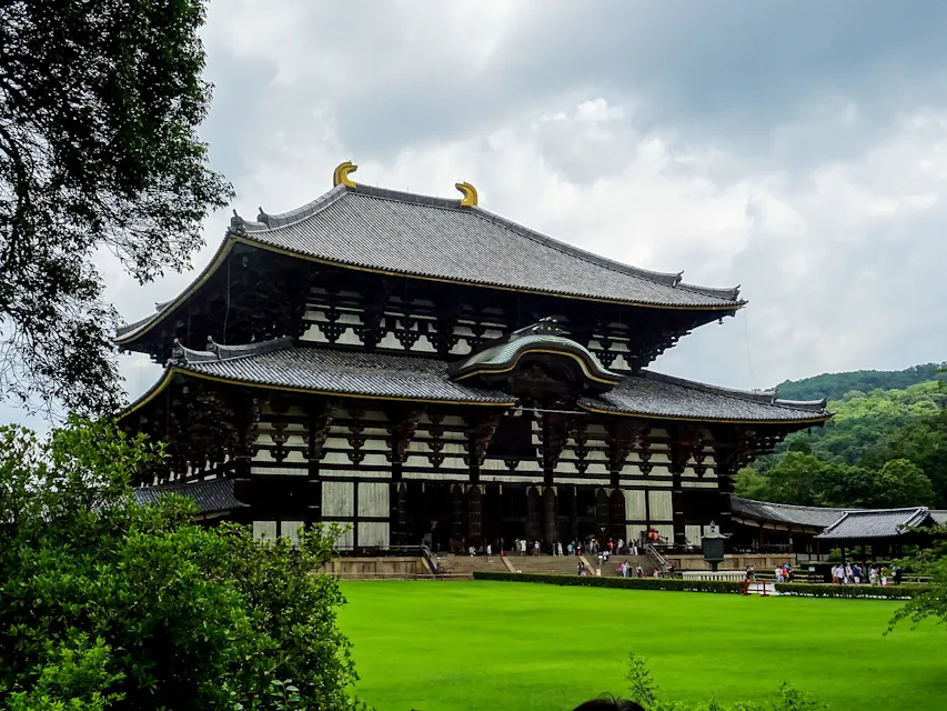 A large, historic temple with traditional Japanese architectural features stands prominently against a cloudy sky. The temple has ornate details and a multi-tiered roof with golden ornaments. A well-manicured lawn and trees surround the structure, and people are gathered at the entrance.