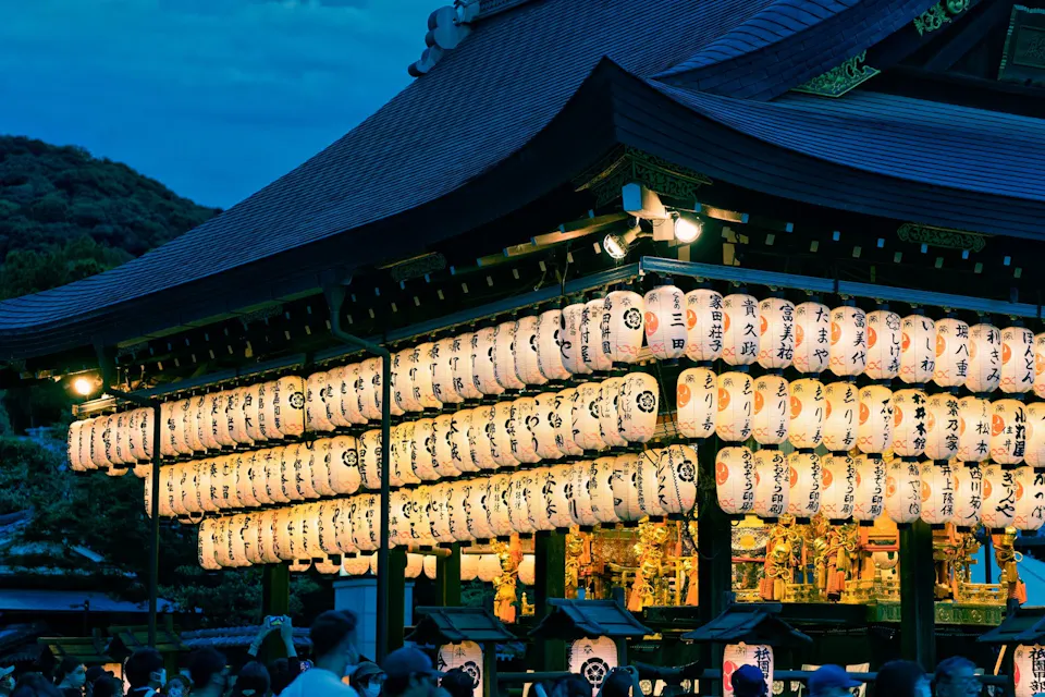 Image of a traditional Japanese festival at dusk. The scene features a beautifully illuminated temple adorned with numerous paper lanterns, each bearing Japanese writing. The lanterns cast a warm glow, and a crowd of people gathers below, enjoying the festive atmosphere.