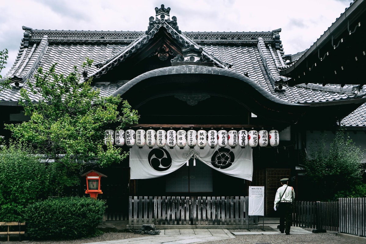 Temple Traditional Japanese temple with ornate roof design and paper lanterns hanging in front. A person in white stands to the right, partially visible. Greenery and a small mailbox-like structure are visible on the left side of the entrance.