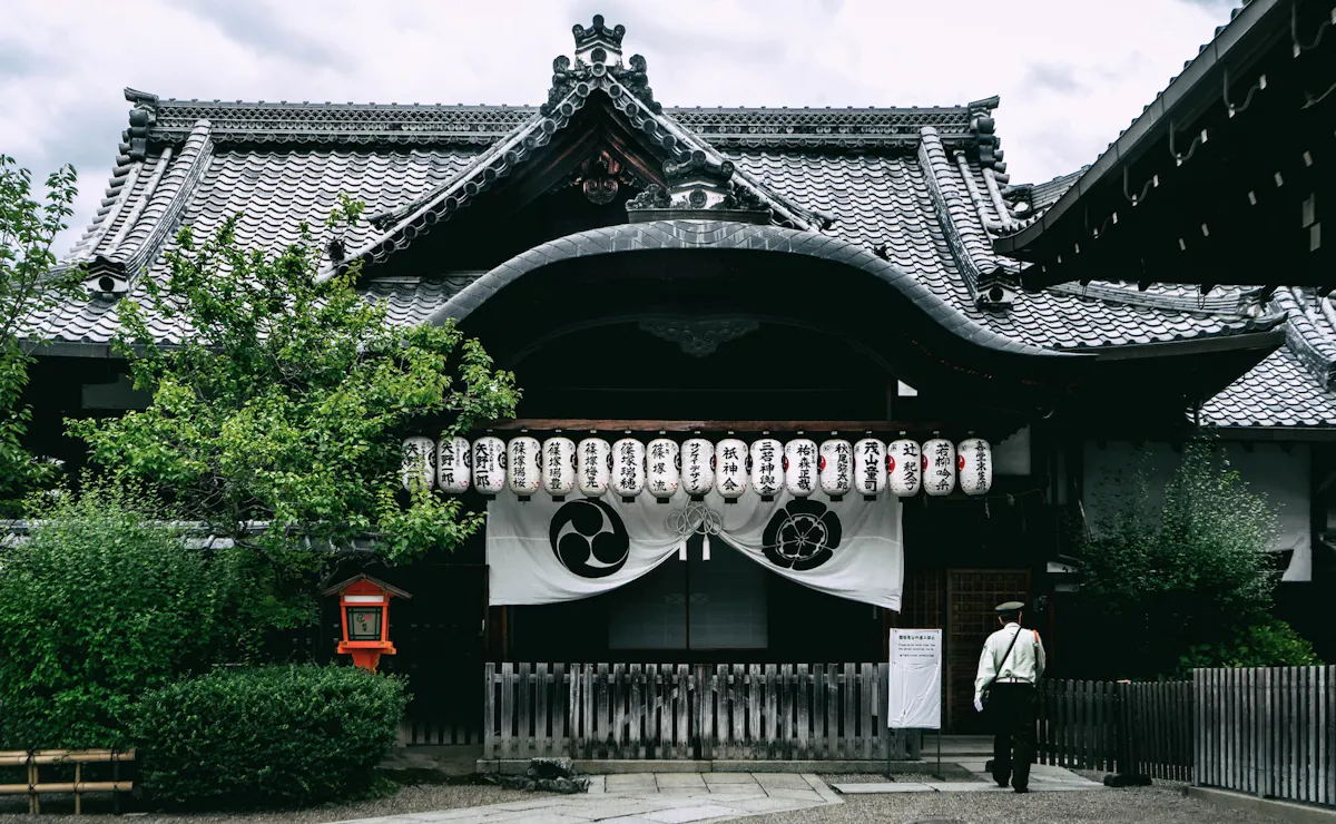 Traditional Japanese temple with ornate roof design and paper lanterns hanging in front. A person in white stands to the right, partially visible. Greenery and a small mailbox-like structure are visible on the left side of the entrance.