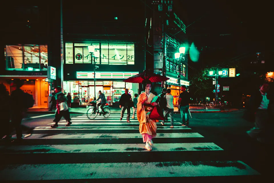 A person wearing traditional Japanese clothing and holding a red parasol walks across a zebra crossing at night. The street is lit with neon signs, and several other people are seen walking and cycling in the background. The atmosphere is vibrant and lively.