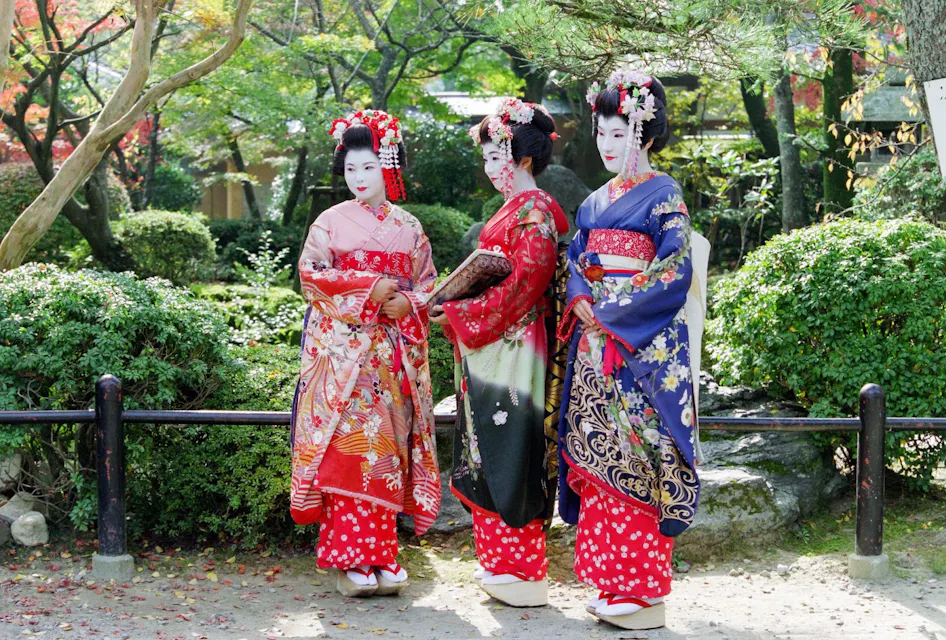 Three women dressed in traditional Geisha attire, with elaborate hairstyles and white face makeup, stand in a garden. The garden features greenery, trees, and a stone pathway. The women are holding fans and wearing ornate kimonos in red, black, and blue.