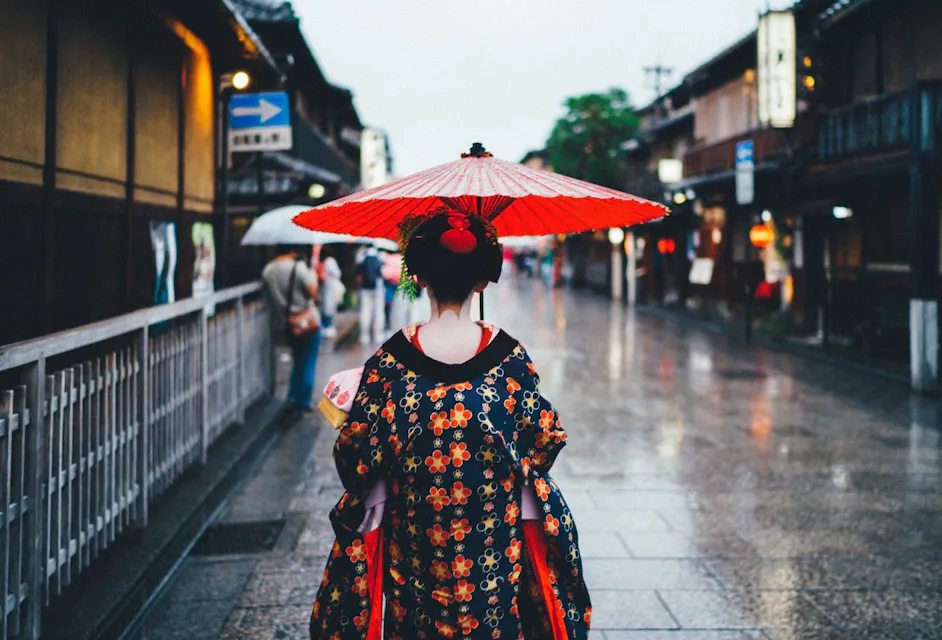 A person wearing a traditional Japanese kimono and holding a red paper umbrella walks down a wet, empty street. The street is lined with wooden buildings, and a few people with umbrellas are visible in the background. The scene appears to be in a historical area.