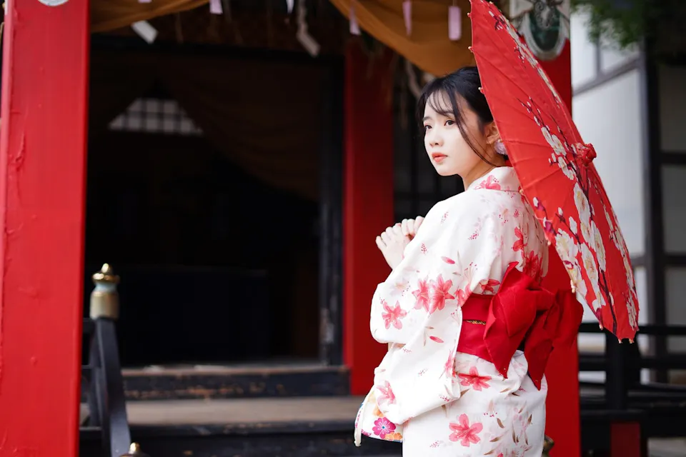 A woman in a floral-patterned kimono holds a red parasol while standing in front of a traditional Japanese building. She gazes to her left, framed by the vibrant red pillars and intricate details of the structure behind her.