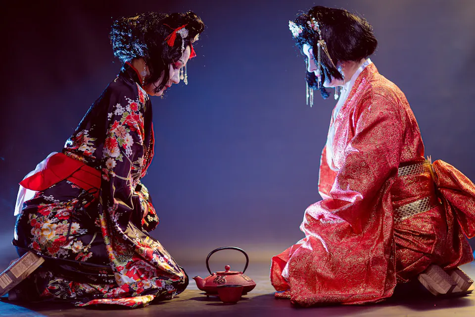 Two individuals dressed in traditional Japanese kimonos kneel facing each other, with a red teapot placed on the ground between them. One kimono is floral patterned with red accents, while the other is a rich, brocade red. Both wear ornate hairpieces.