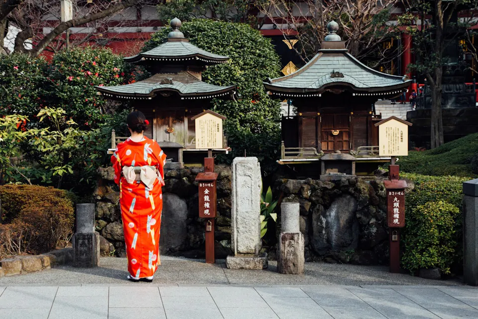 A person wearing a traditional red kimono stands in front of a small shrine surrounded by lush plants and trees. The shrine features wooden structures with green-tiled roofs and informational placards.