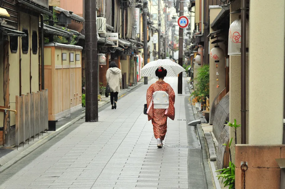 A person wearing a traditional Japanese kimono and holding a white umbrella walks down a narrow, quiet street lined with old wooden buildings. Another individual walks ahead in the distance on the same street.