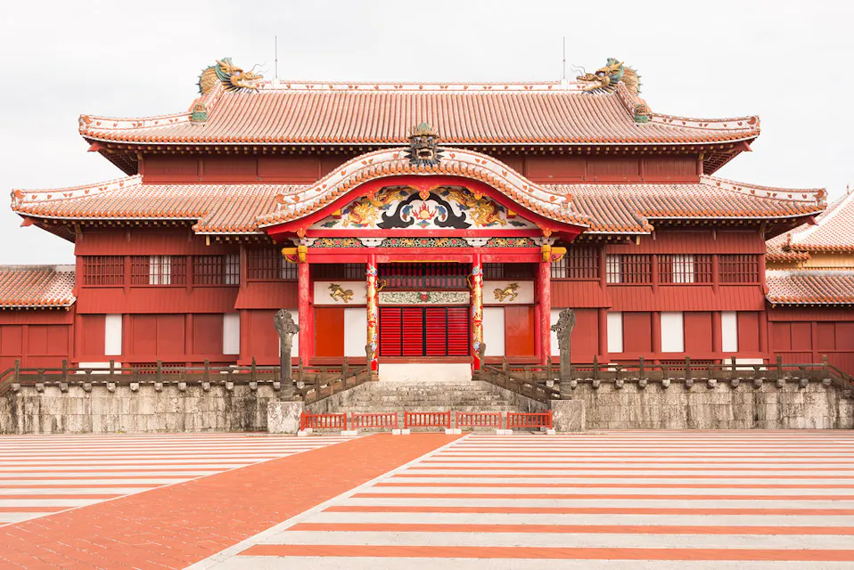 Nakagusuku Castle Ruins 一张日本冲绳首里城的照片,展示了其鲜艳的红色和传统的琉球建筑,装饰华丽,拥有一扇大红门和复杂的屋顶细节。前景呈现红白相间的砖砌。