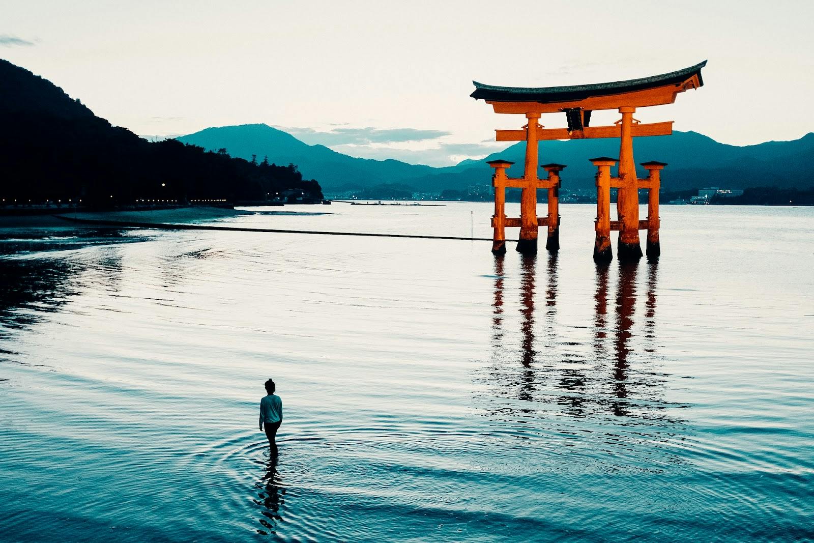 A person stands in shallow water at sunset, facing a large, traditional red torii gate rising from the water, with mountains and a distant shoreline in the background.