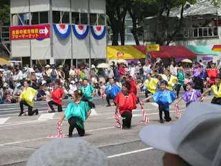 A lively outdoor parade with dancers in colorful outfits performing in formation on a street, holding striped sticks. Crowds watch from behind barriers, with booths and banners visible in the background.