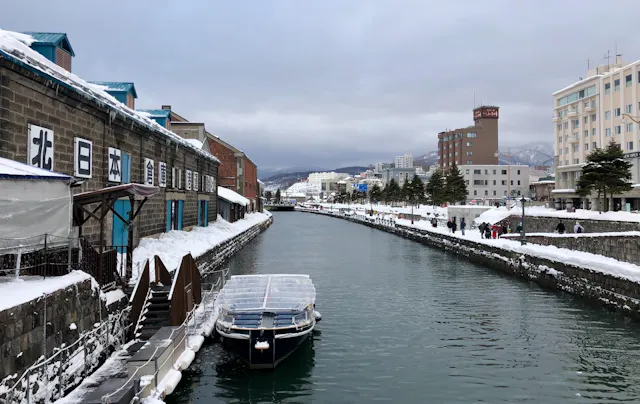 A snowy canal scene with a covered boat docked along historic brick warehouses on the left, and modern buildings on the right, under a cloudy sky. Snow blankets the rooftops and ground.
