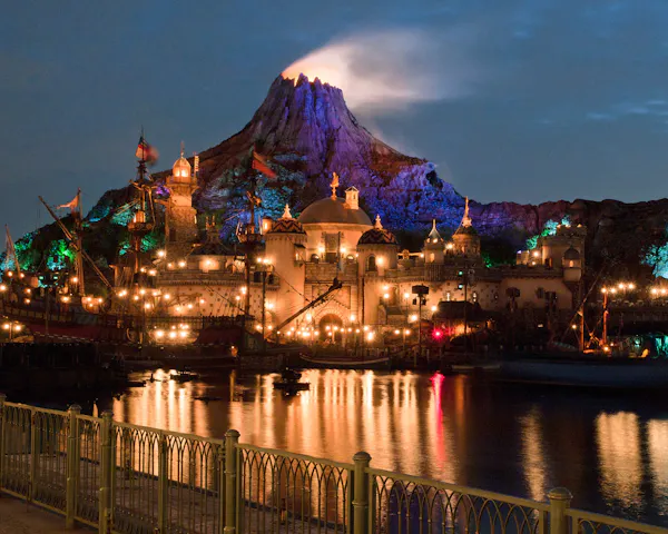 A glowing volcano rises behind an illuminated theme park at night, with colorful lights reflecting on the calm water in the foreground and ornate buildings creating a magical atmosphere.