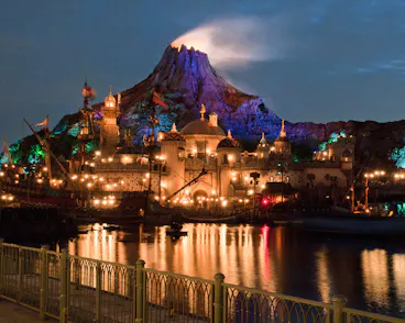 A glowing volcano rises behind an illuminated theme park at night, with colorful lights reflecting on the calm water in the foreground and ornate buildings creating a magical atmosphere.