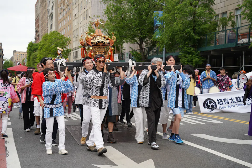 A lively group of people in traditional clothing carry a decorative portable shrine during a city street parade. They are surrounded by participants in colorful attire and spectators, with trees and buildings in the background.