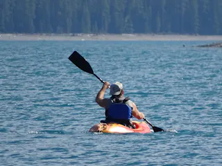 A person wearing a life jacket and cap paddles a red kayak on a calm blue lake surrounded by forested shoreline in the background.