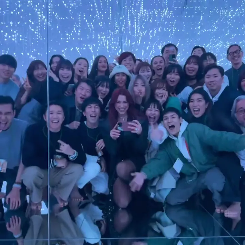 A large group of people, energetically posing for a photo in front of a mirror, surrounded by a starry, illuminated backdrop. One person in the center is taking the photo with a smartphone. The atmosphere appears joyful and lively.