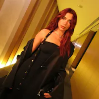 A person with long reddish hair stands in a hallway with golden lighting. They are wearing a black strapless top and black button-up jacket that is draped off one shoulder. The hallway has vertical paneling and recessed lighting along the walls.