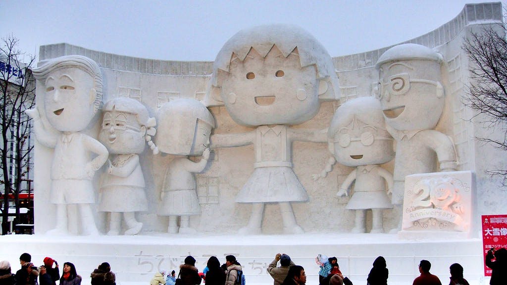 Large snow sculpture of five cartoon characters, including one in the center pointing, with a crowd of people in winter clothing standing and walking in front. A sign celebrating a 20th anniversary is on the right.
