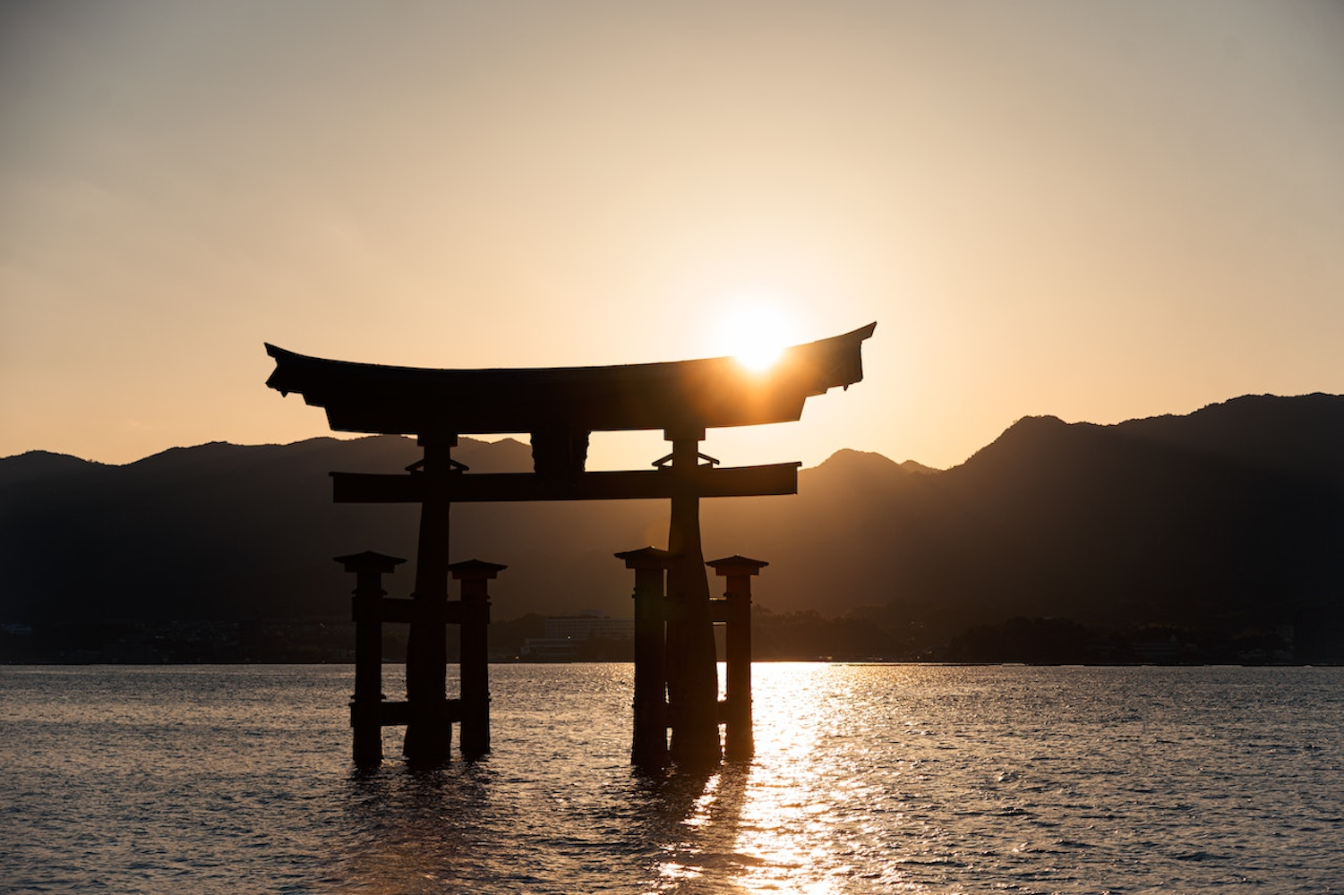 Silhouette of a traditional Japanese torii gate standing in the water with the sun setting behind it. The sky is a warm gradient of golden hues, and dark mountains are visible in the background. The water reflects the light from the setting sun.