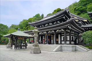 A traditional Japanese temple with ornate black-and-white wooden architecture, a green-roofed pavilion, stone lanterns, and lush greenery in the background. Several people are near the entrance and on the temple steps.