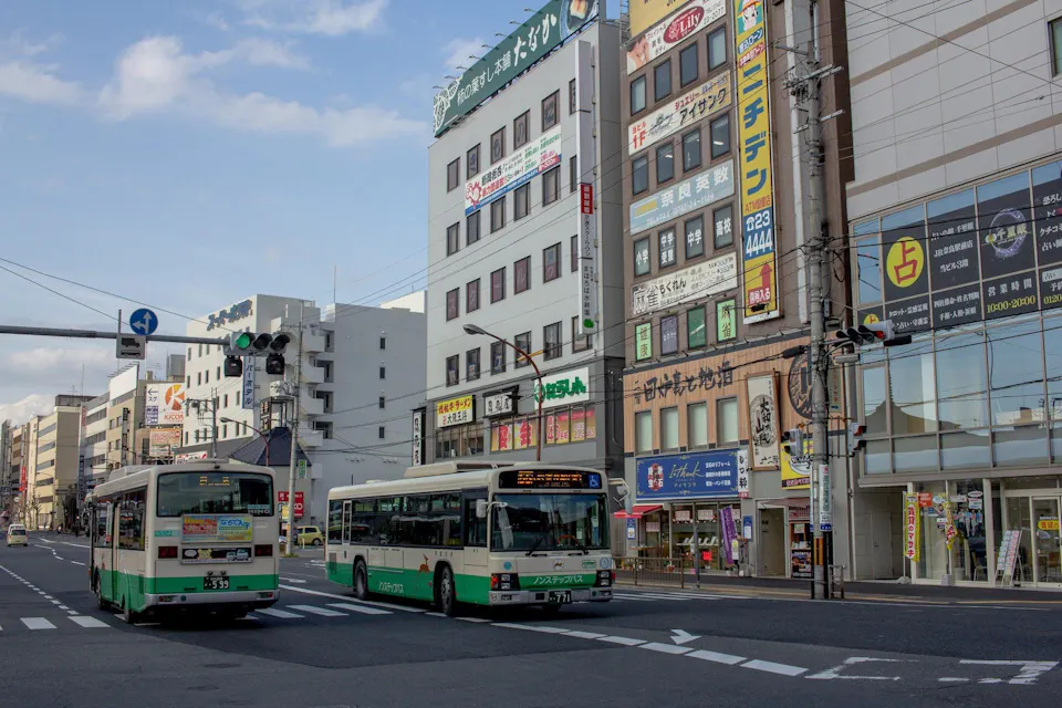 Buses in Japan A busy urban street with two green and white buses at a traffic intersection. Several multi-story buildings with numerous signs and advertisements in Japanese line the street. Pedestrians can be seen walking on the sidewalks. The sky is partly cloudy.