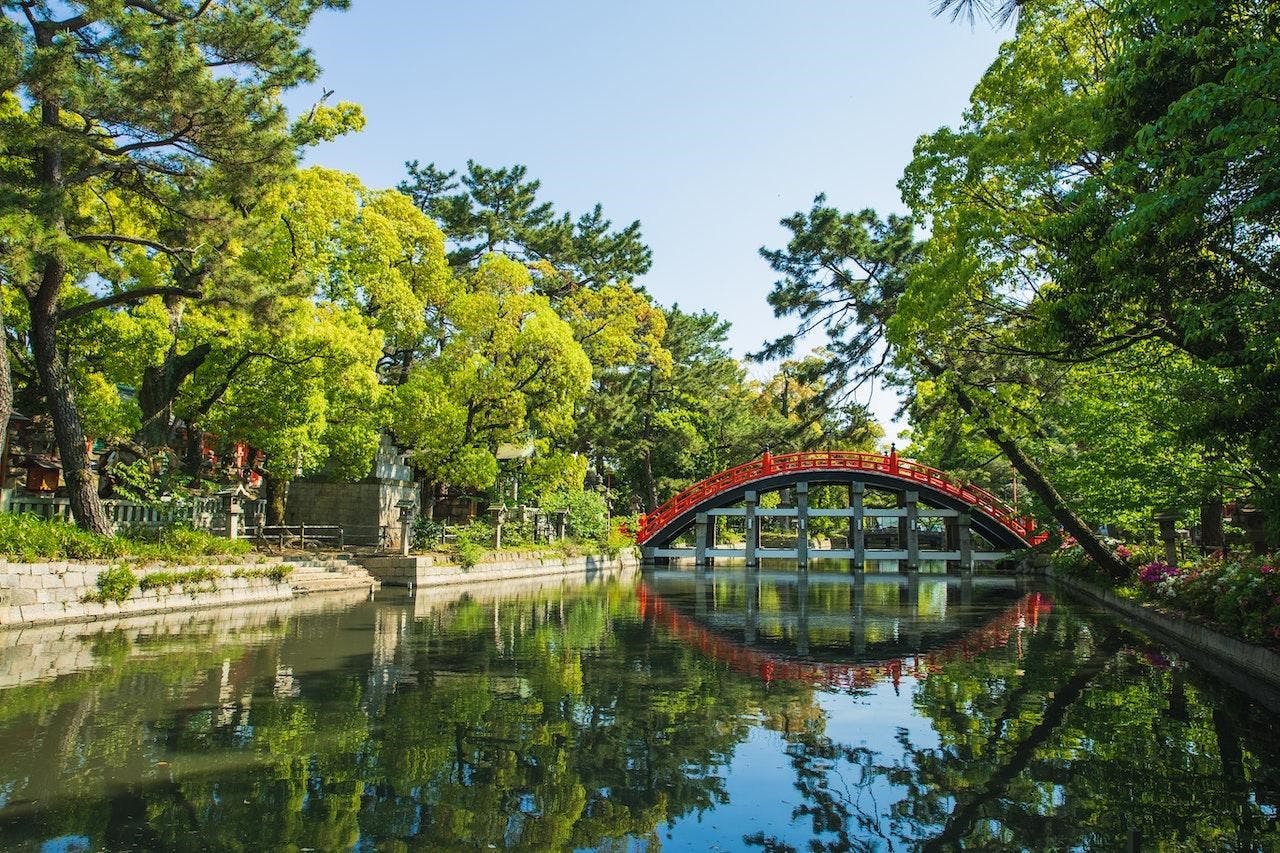 A vibrant red arched bridge crosses over a serene pond surrounded by lush green trees and foliage under a clear blue sky, with reflections visible in the calm water.