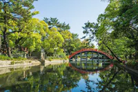 A vibrant red arched bridge crosses over a serene pond surrounded by lush green trees and foliage under a clear blue sky, with reflections visible in the calm water.