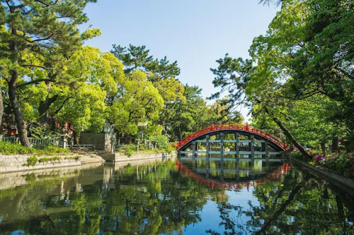 A vibrant red arched bridge crosses over a serene pond surrounded by lush green trees and foliage under a clear blue sky, with reflections visible in the calm water.