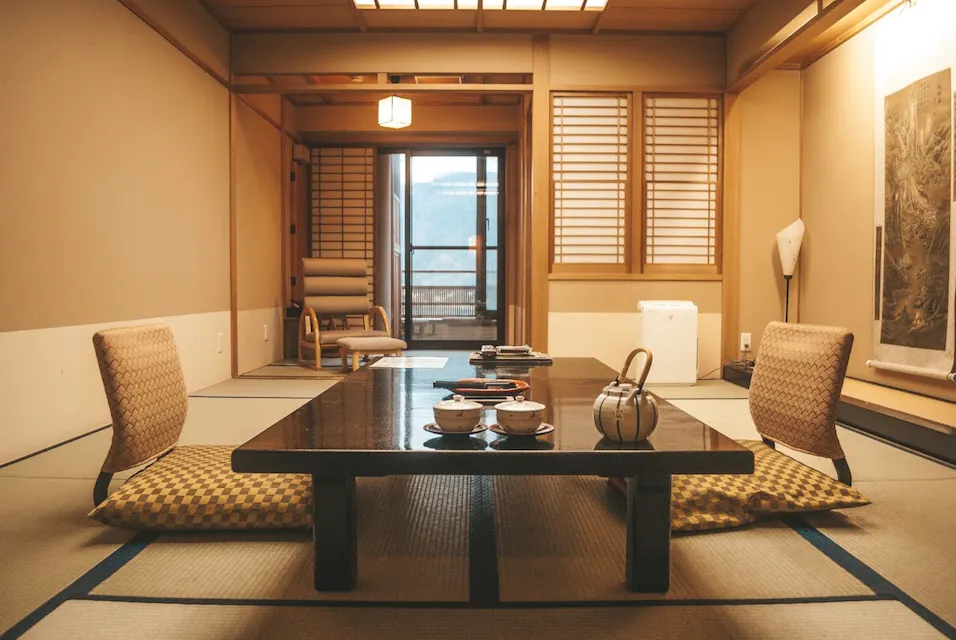 A traditional Japanese room with tatami mats, a low wooden table set for tea, and two cushioned floor seats. Shoji screens cover the windows, and a scroll hangs on the wall. A small chair is visible near the door leading to a balcony.