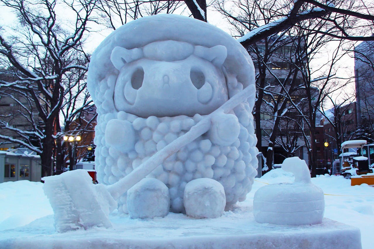 A large, detailed snow sculpture of a cute, cartoon-like sheep holding a broom, sitting outdoors in a snowy park with bare trees and buildings in the background at dusk.