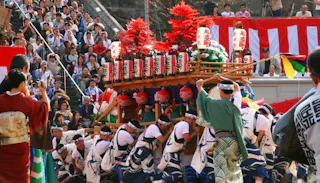 A group of people in traditional Japanese clothing carry a colorful festival float with red decorations and lanterns, while a crowd watches the lively scene outdoors.