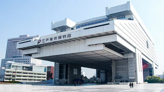 A modern, large, white building with a unique elevated design supported by pillars. The sign in Japanese reads "Edo-Tokyo Museum." People are visible walking in the plaza below on a clear day.