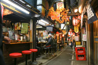 A narrow alley in Japan is adorned with lanterns and hanging autumn leaves, bustling with small eateries. People are seated on stools at a bar counter, enjoying meals. Menus in Japanese are displayed, and crates of Sapporo beer are visible in the foreground.