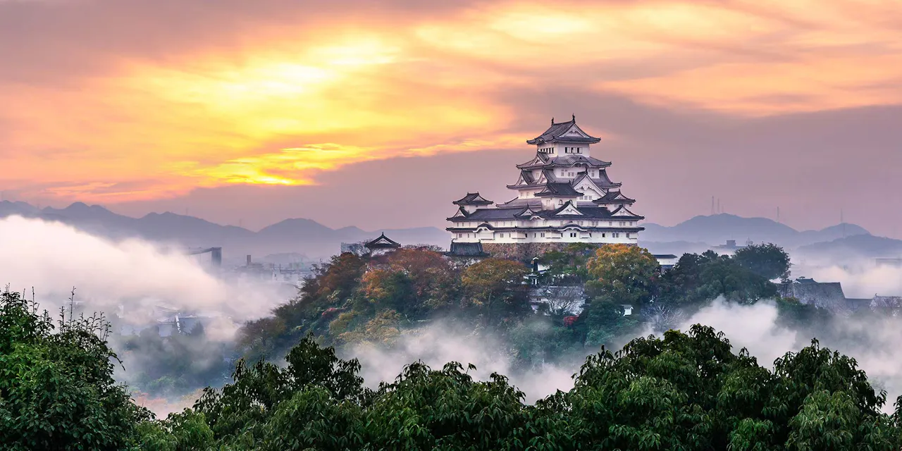 A majestic Japanese castle, Himeji Castle, stands atop a hill surrounded by fog and lush greenery. The sky is painted with hues of orange and yellow from the setting sun, casting a serene glow over the landscape. The distant mountains add depth to the picturesque scene.