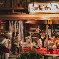 Nagoya Food Stall An evening scene of a bustling Japanese izakaya. Patrons in casual attire are gathered around tables, enjoying food and drinks. The izakaya's name is displayed on a sign above, and traditional Japanese decor and lighting add to the warm ambiance.