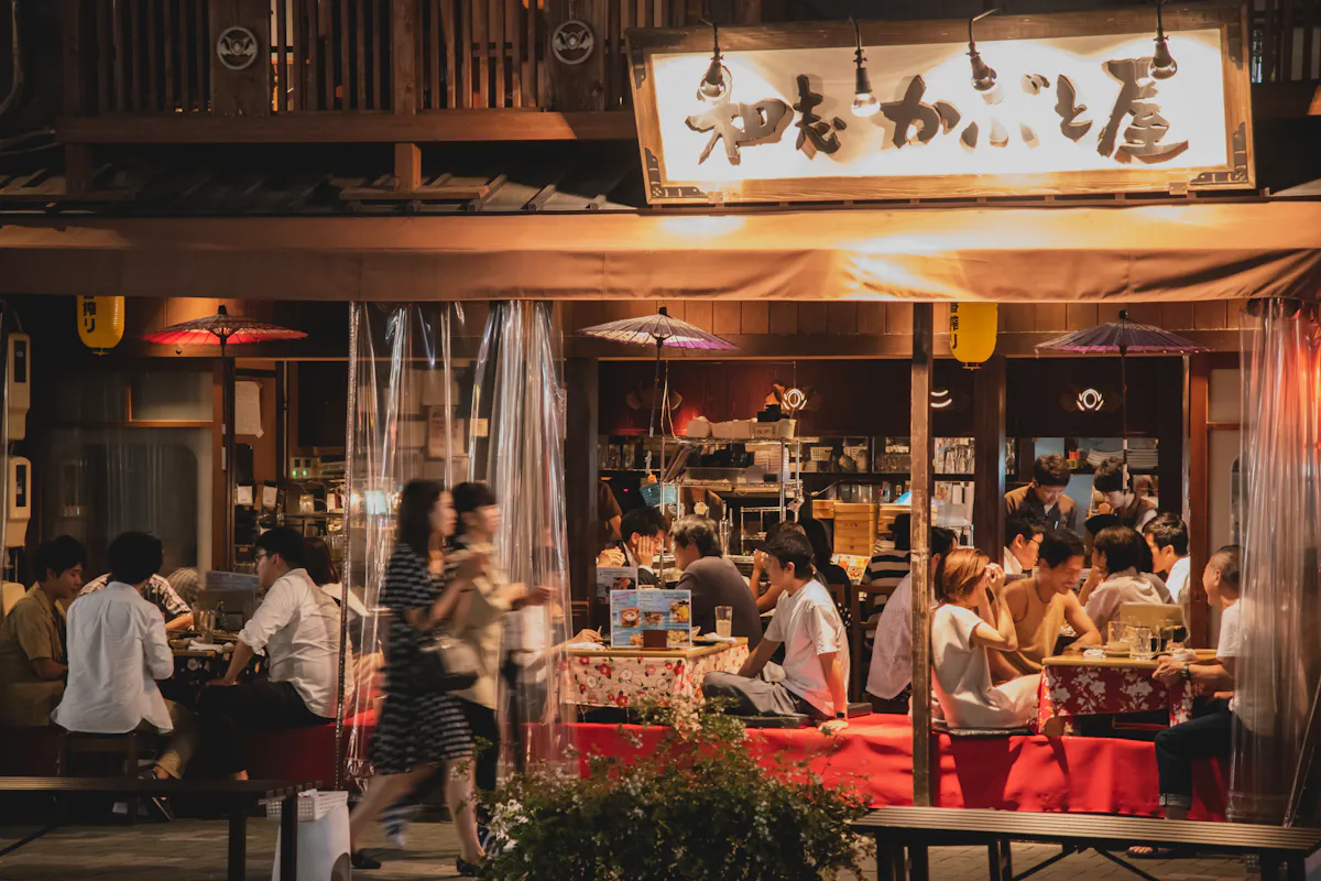 Nagoya Food Stall An evening scene of a bustling Japanese izakaya. Patrons in casual attire are gathered around tables, enjoying food and drinks. The izakaya's name is displayed on a sign above, and traditional Japanese decor and lighting add to the warm ambiance.