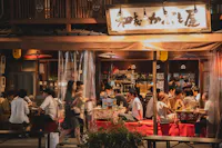 An evening scene of a bustling Japanese izakaya. Patrons in casual attire are gathered around tables, enjoying food and drinks. The izakaya's name is displayed on a sign above, and traditional Japanese decor and lighting add to the warm ambiance.