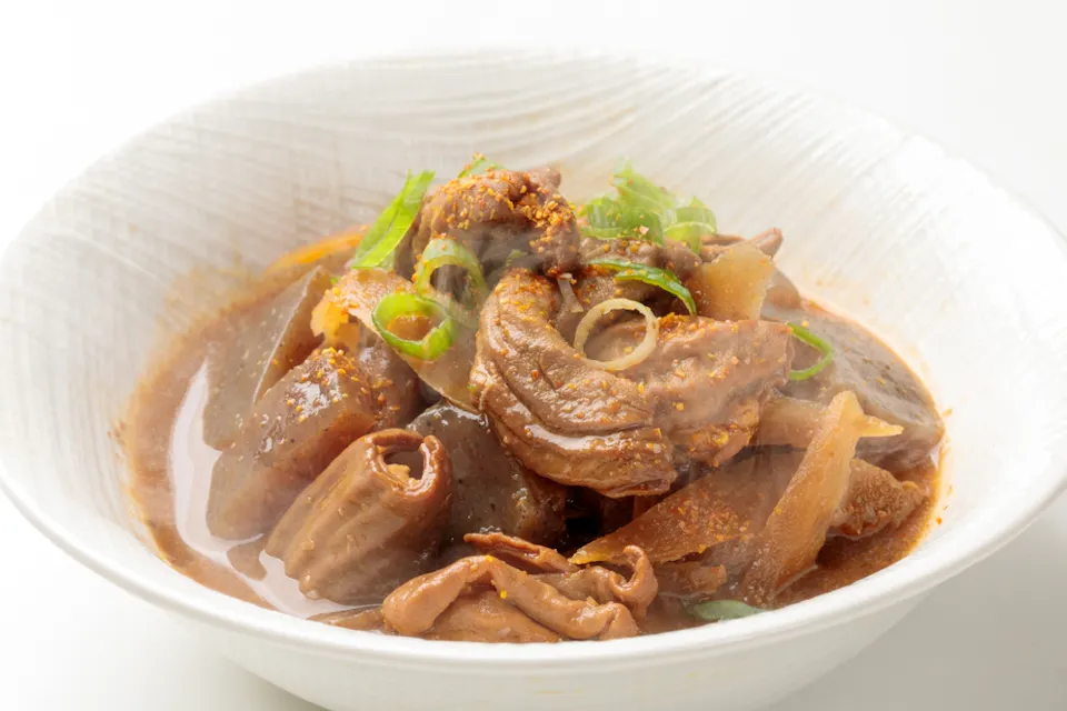 A close-up of a white bowl filled with a savory stew, featuring tender pieces of meat and intestines, garnished with chopped green onions and a sprinkling of seasoning. The broth has a rich, dark color, and steam rises from the hot dish.