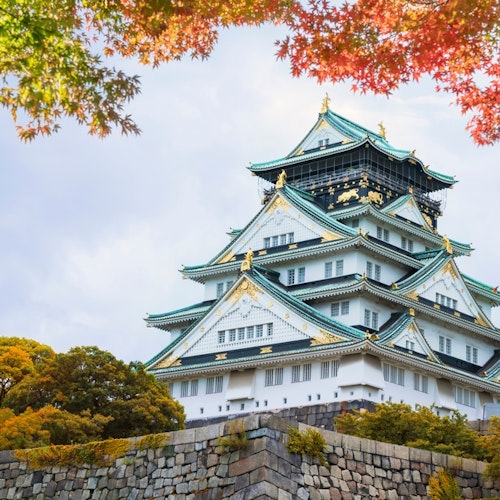 Osaka Castle A traditional Japanese castle with white walls and teal roofs sits atop a stone base, surrounded by vibrant foliage in shades of green, yellow, and red. The sky above is mostly cloudy with hints of blue peeking through.