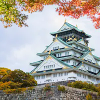 Osaka Castle A traditional Japanese castle with white walls and teal roofs sits atop a stone base, surrounded by vibrant foliage in shades of green, yellow, and red. The sky above is mostly cloudy with hints of blue peeking through.